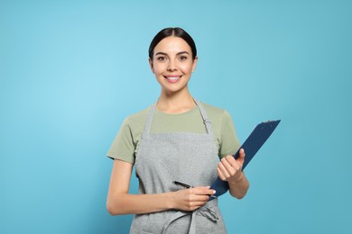 Young woman in grey apron with clipboard on light blue background