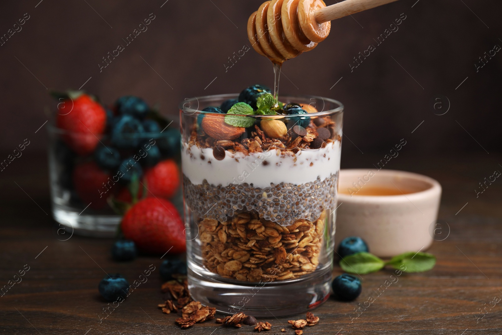 Photo of Pouring honey onto tasty granola with berries, yogurt and chia seeds at wooden table, closeup