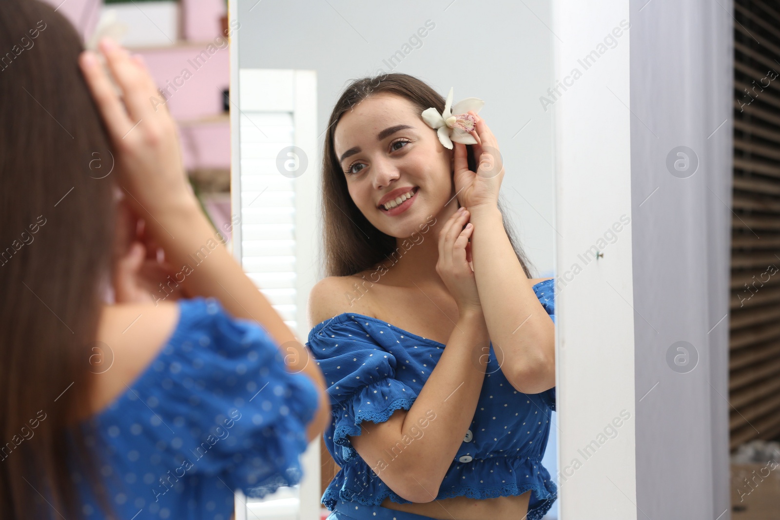 Photo of Beautiful young woman with orchid flower looking at herself in large mirror indoors