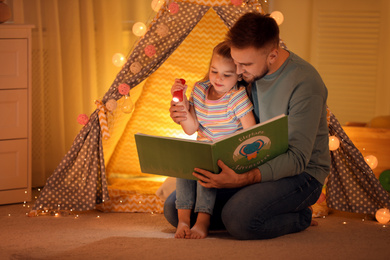 Photo of Father and daughter with flashlight reading book at home