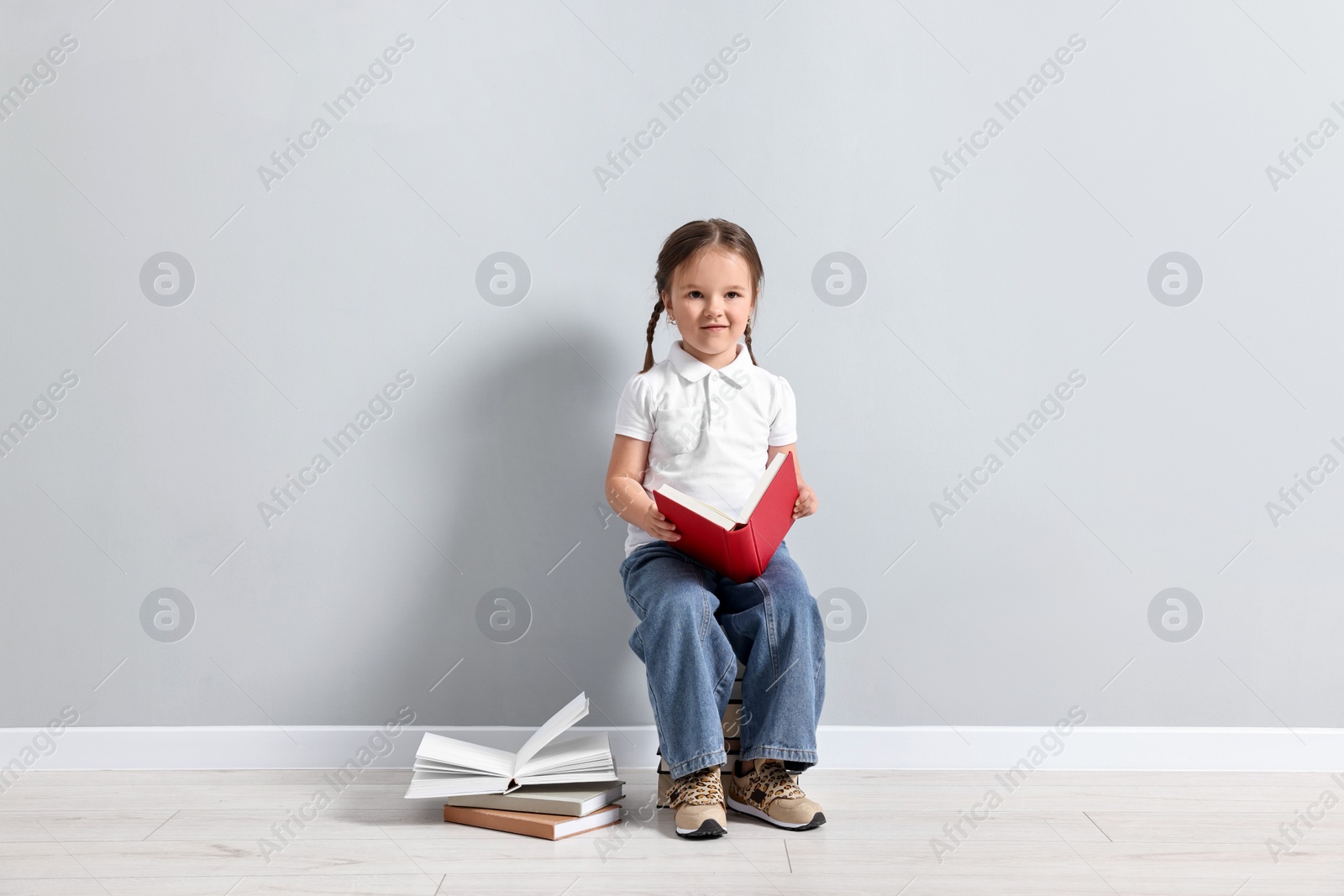 Photo of Cute little girl sitting on stack of books near light grey wall