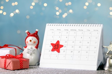 Saint Nicholas Day. Calendar with marked date December 19, gift boxes and snowman figure on table against blurred lights
