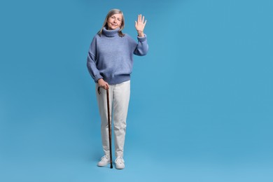 Senior woman with walking cane waving on light blue background