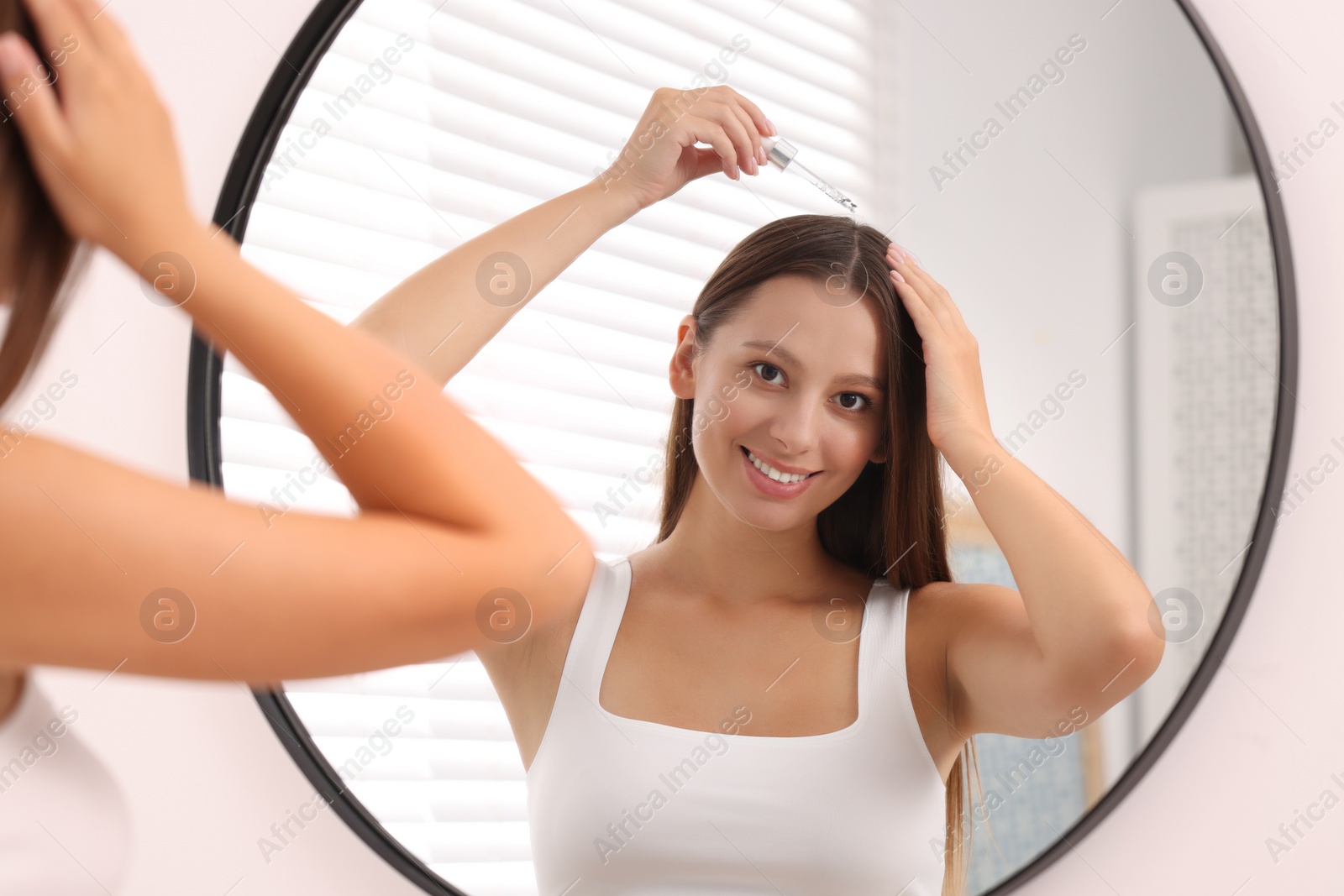 Photo of Beautiful woman applying serum onto hair near mirror indoors