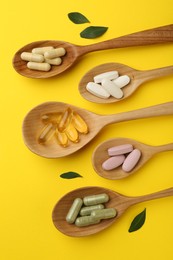 Photo of Different vitamin pills in spoons and green leaves on yellow background, flat lay