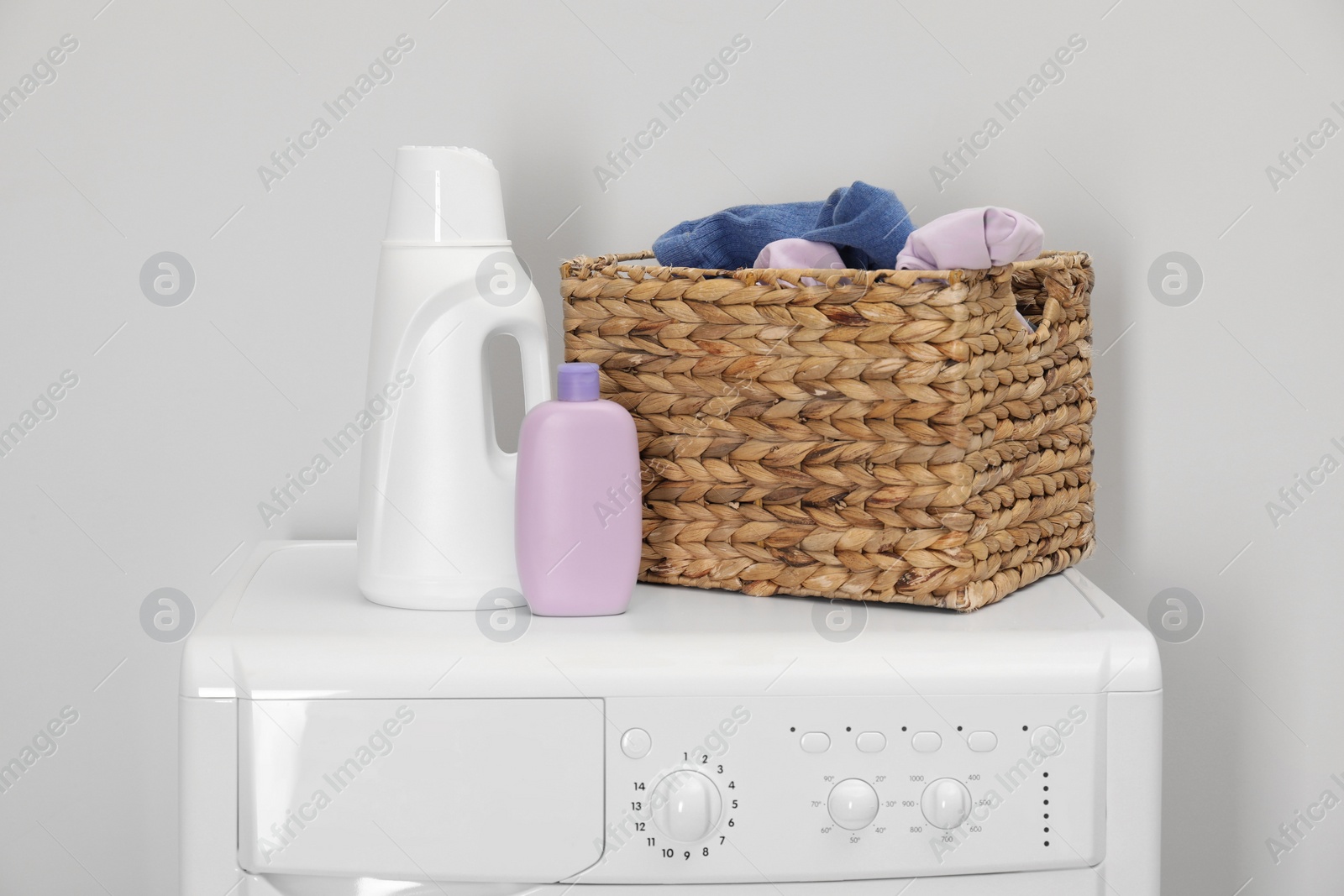 Photo of Baby clothes in wicker basket and laundry detergents on washing machine near light wall