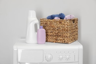 Baby clothes in wicker basket and laundry detergents on washing machine near light wall