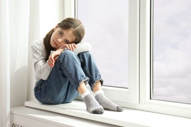 Photo of Sad little girl sitting on window sill indoors, space for text
