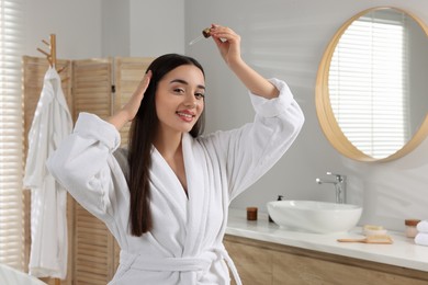 Photo of Happy young woman applying essential oil onto hair roots in bathroom