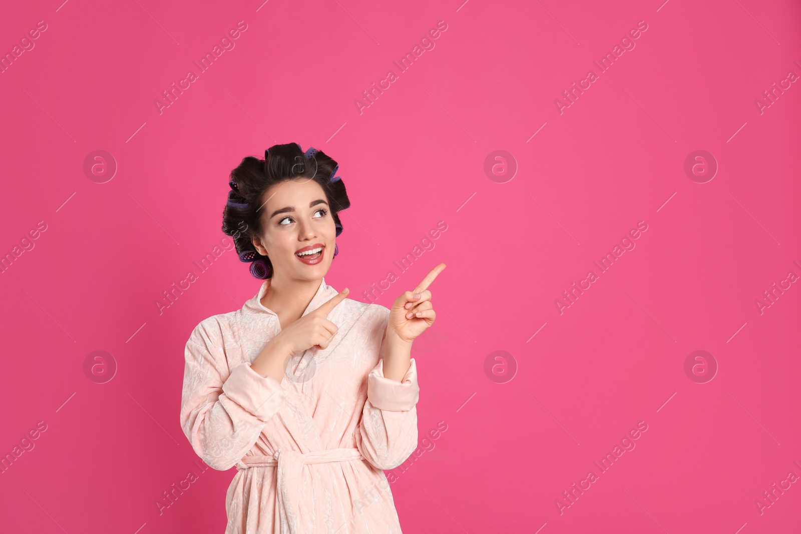 Photo of Happy young woman in bathrobe with hair curlers on pink background, space for text