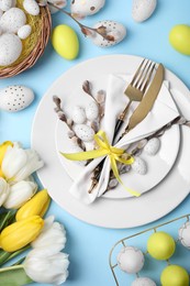 Photo of Festive table setting with tulips and painted eggs on light blue background, flat lay. Easter celebration