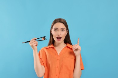 Photo of Emotional young woman holding sushi roll with chopsticks on light blue background