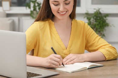 Photo of Woman writing in notebook while working on laptop at wooden table indoors