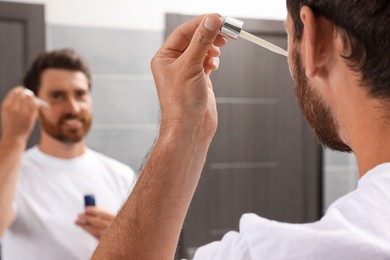 Photo of Handsome man applying oil on beard in bathroom, closeup