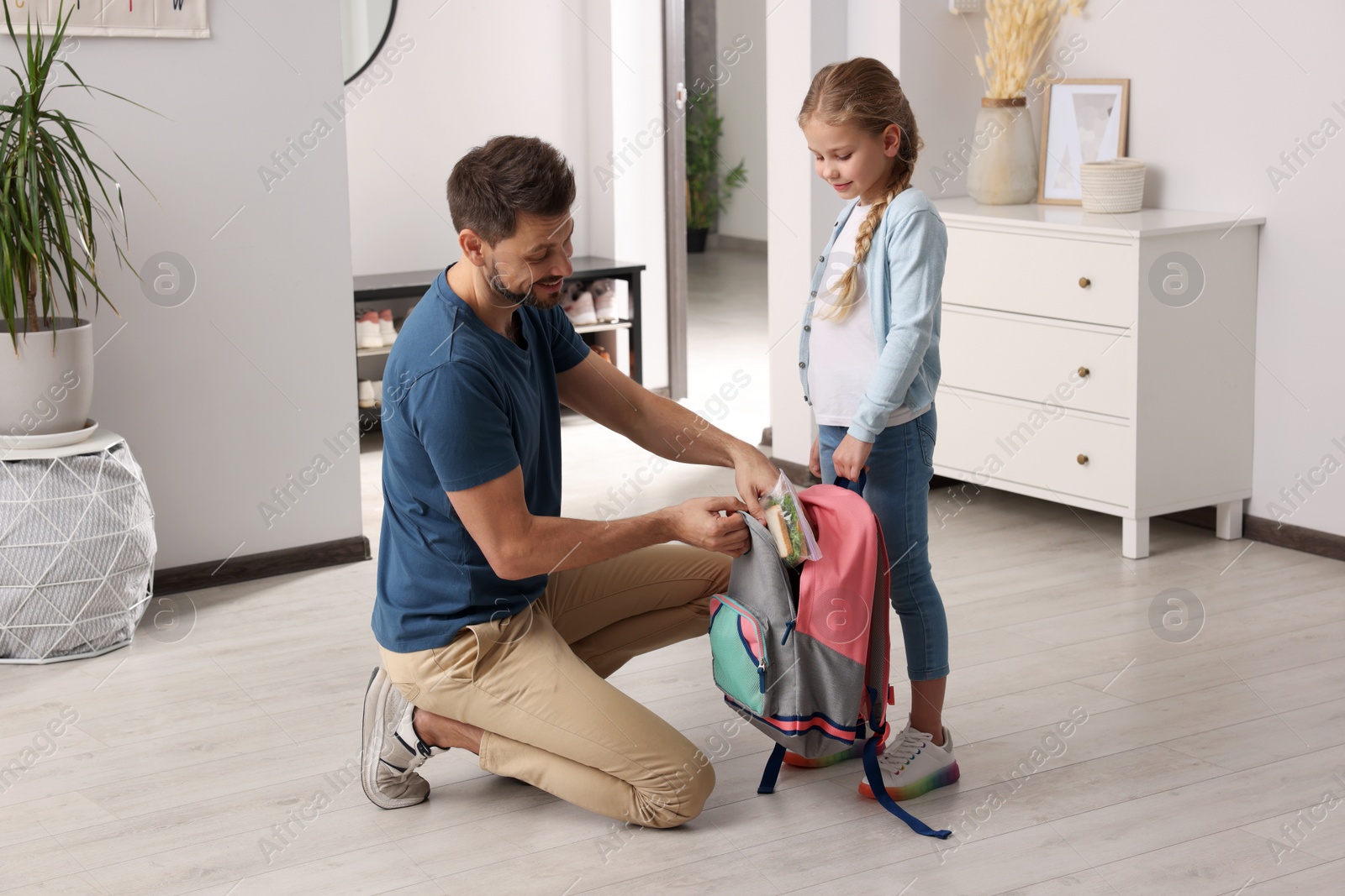 Photo of Happy father putting lunch box into daughter`s backpack at home. Preparing to school