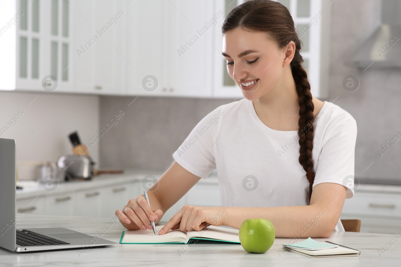Photo of Beautiful young woman writing in notebook while working on laptop at white marble table indoors