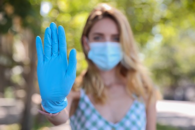Photo of Woman in protective face mask showing stop gesture outdoors, focus on hand. Prevent spreading of coronavirus