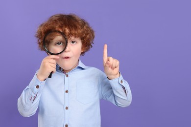 Photo of Cute little boy looking through magnifier glass and pointing on violet background, space for text