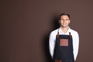 Photo of Portrait of happy young waiter in uniform on brown background, space for text