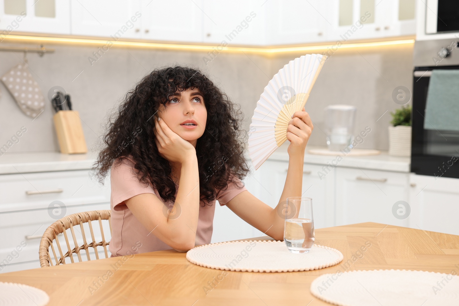 Photo of Young woman waving hand fan to cool herself at table in kitchen