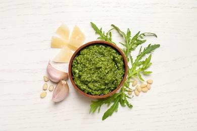 Bowl of tasty arugula pesto and ingredients on white wooden table, flat lay