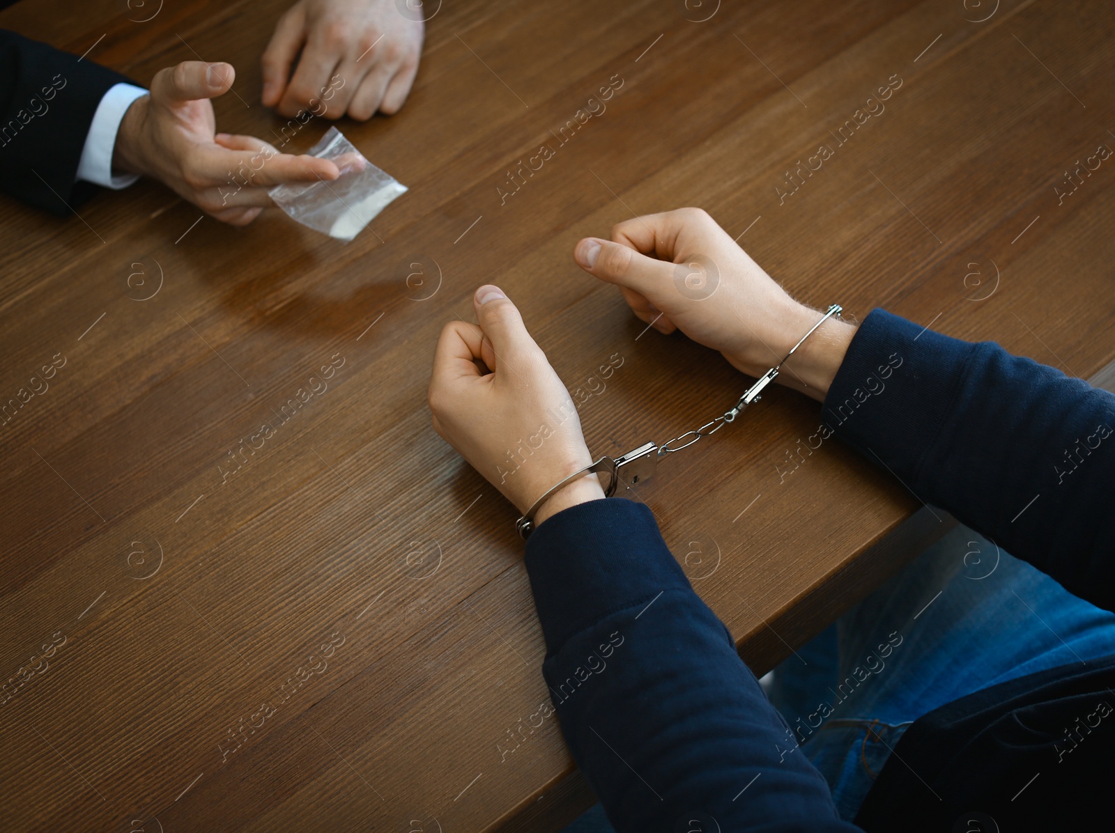 Photo of Police officer interrogating drug dealer in handcuffs at desk indoors. Criminal law