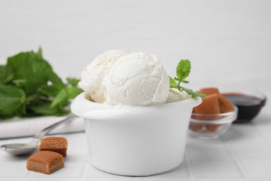 Photo of Scoops of tasty ice cream with mint leaves and caramel candies on white tiled table, closeup