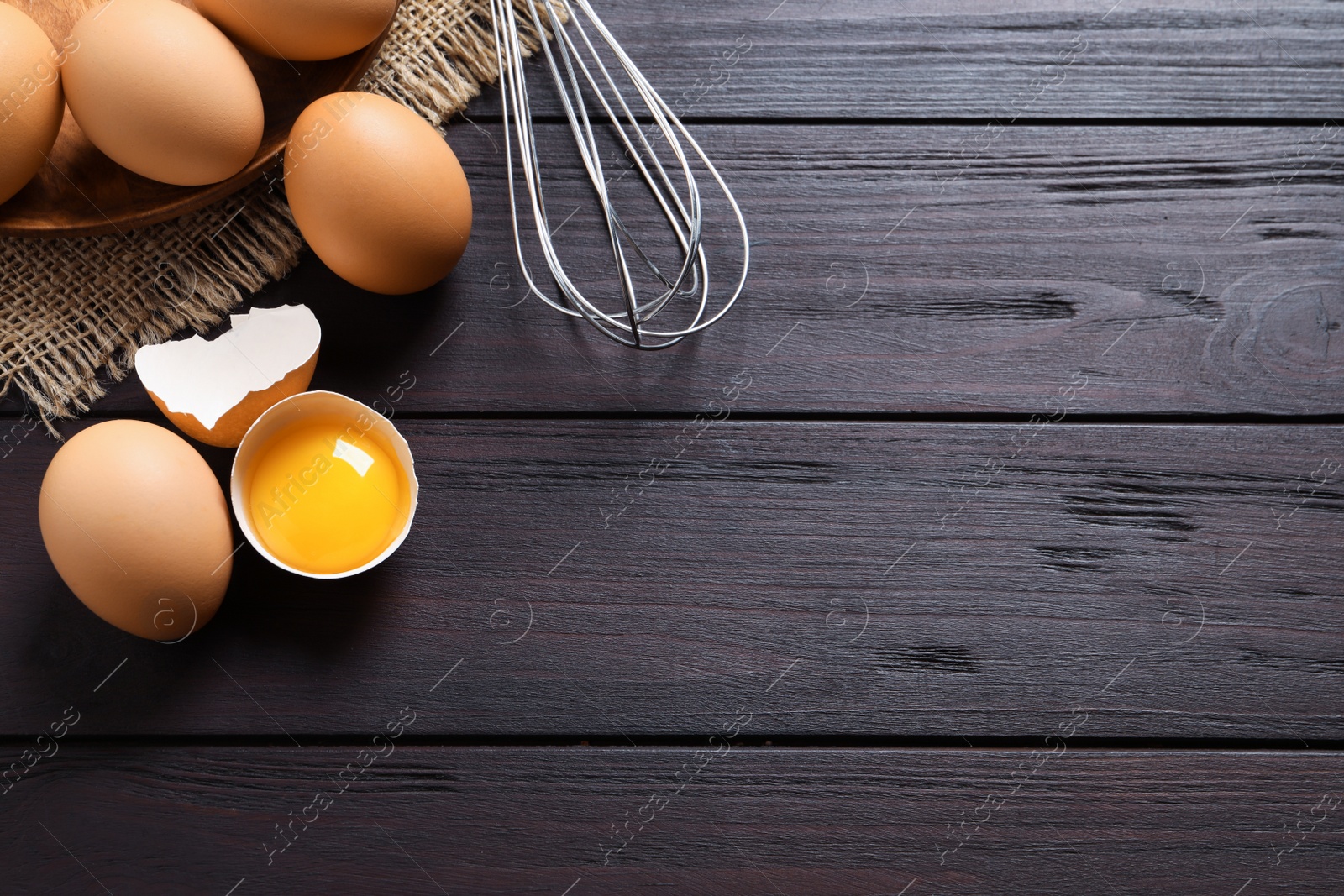 Photo of Raw chicken eggs, shell with yolk and whisk on dark wooden table, flat lay. Space for text