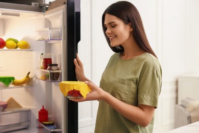 Photo of Woman holding bowl of fresh tomatoes covered with beeswax food wrap near refrigerator in kitchen