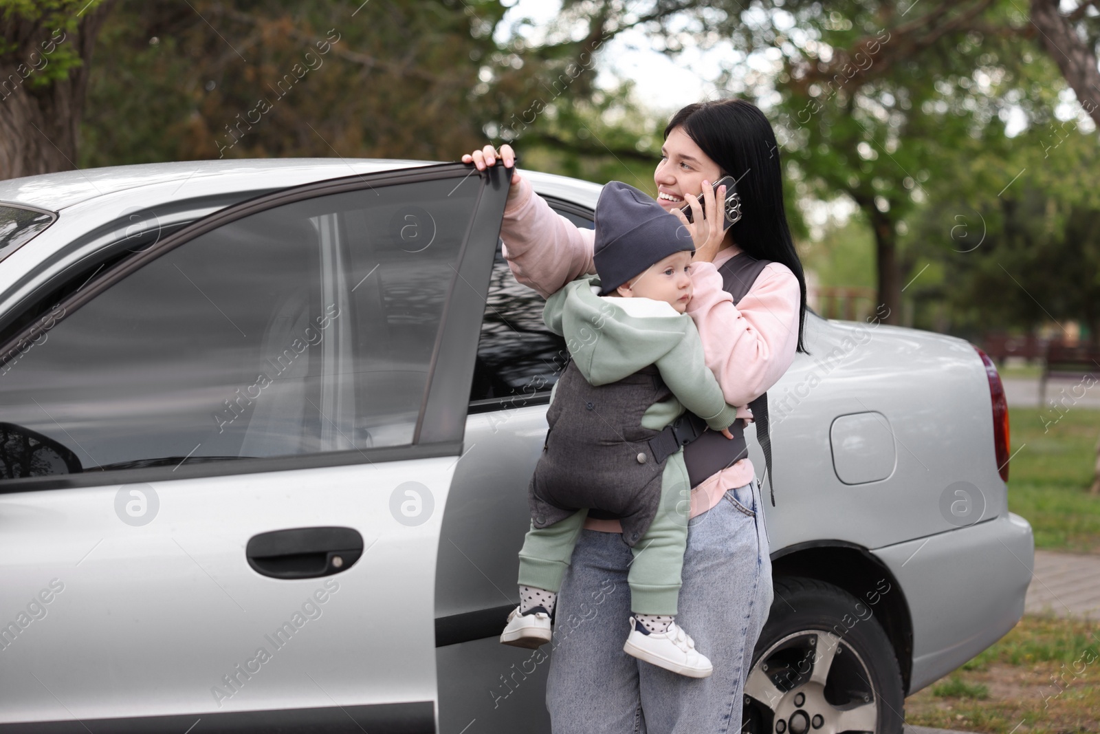 Photo of Mother holding her child in sling (baby carrier) while talking on smartphone near car outdoors