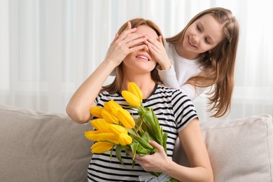 Daughter covering mother's eyes with her palms and congratulating with bouquet of yellow tulips at home
