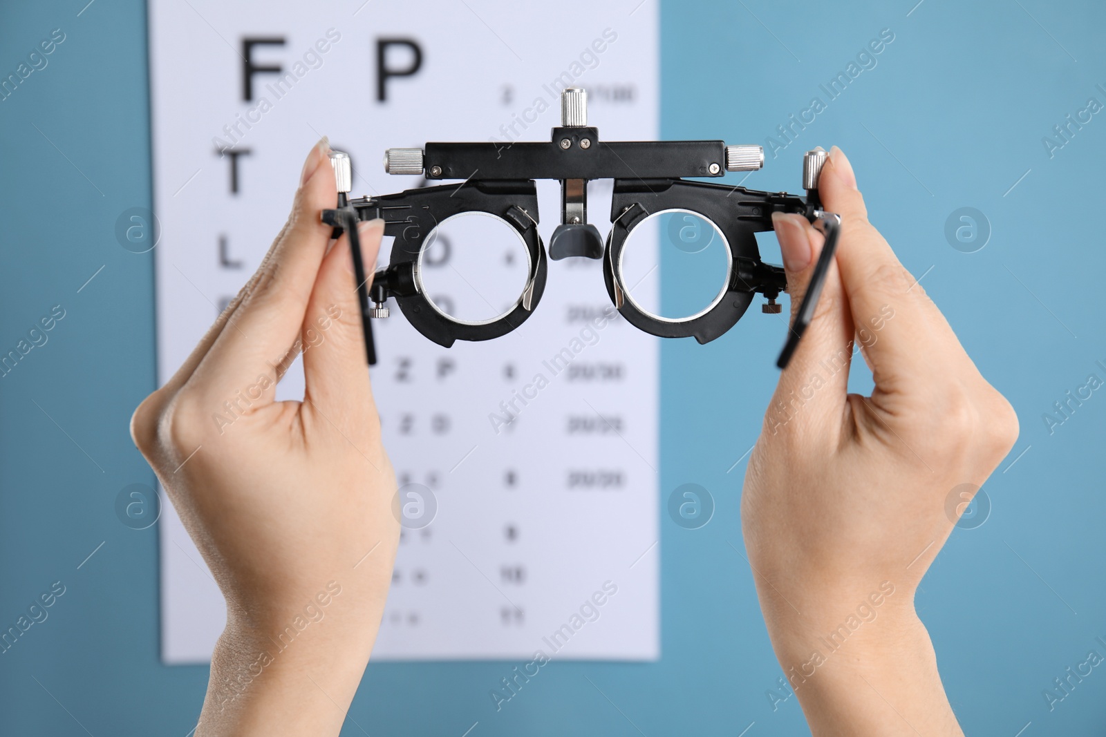 Photo of Woman holding trial frame against eye chart test on blue background, closeup. Ophthalmologist tool