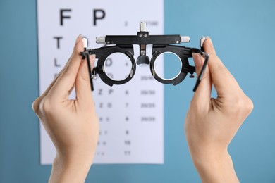 Woman holding trial frame against eye chart test on blue background, closeup. Ophthalmologist tool