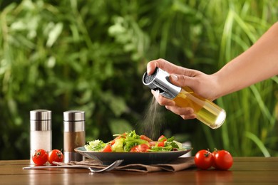 Photo of Woman spraying cooking oil onto delicious salad at wooden table against blurred green background, closeup