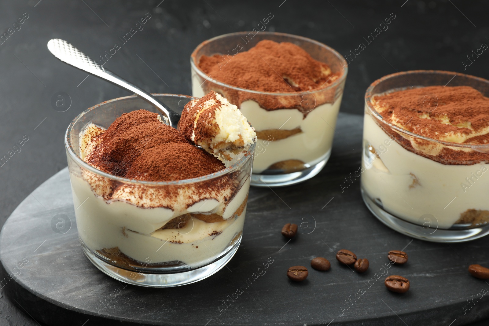 Photo of Delicious tiramisu in glasses, spoon and coffee beans on black table