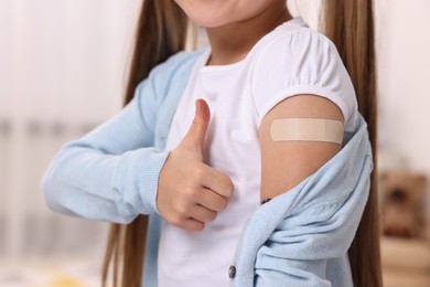 Photo of Girl with sticking plaster on arm after vaccination showing thumbs up indoors, closeup