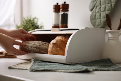 Photo of Woman taking loaf from wooden bread basket at white marble table in kitchen, closeup