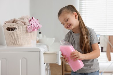 Photo of Little girl pouring fabric softener into washing machine in bathroom