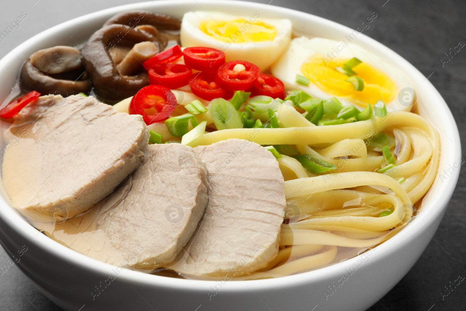 Photo of Delicious ramen with meat and eggs in bowl on grey textured table, closeup. Noodle soup