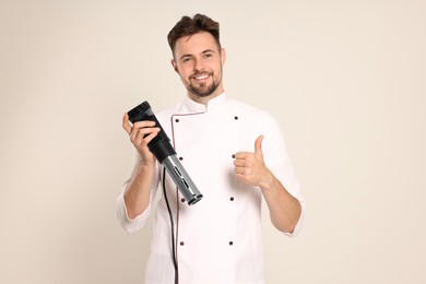Photo of Smiling chef holding sous vide cooker and showing thumb up on beige background