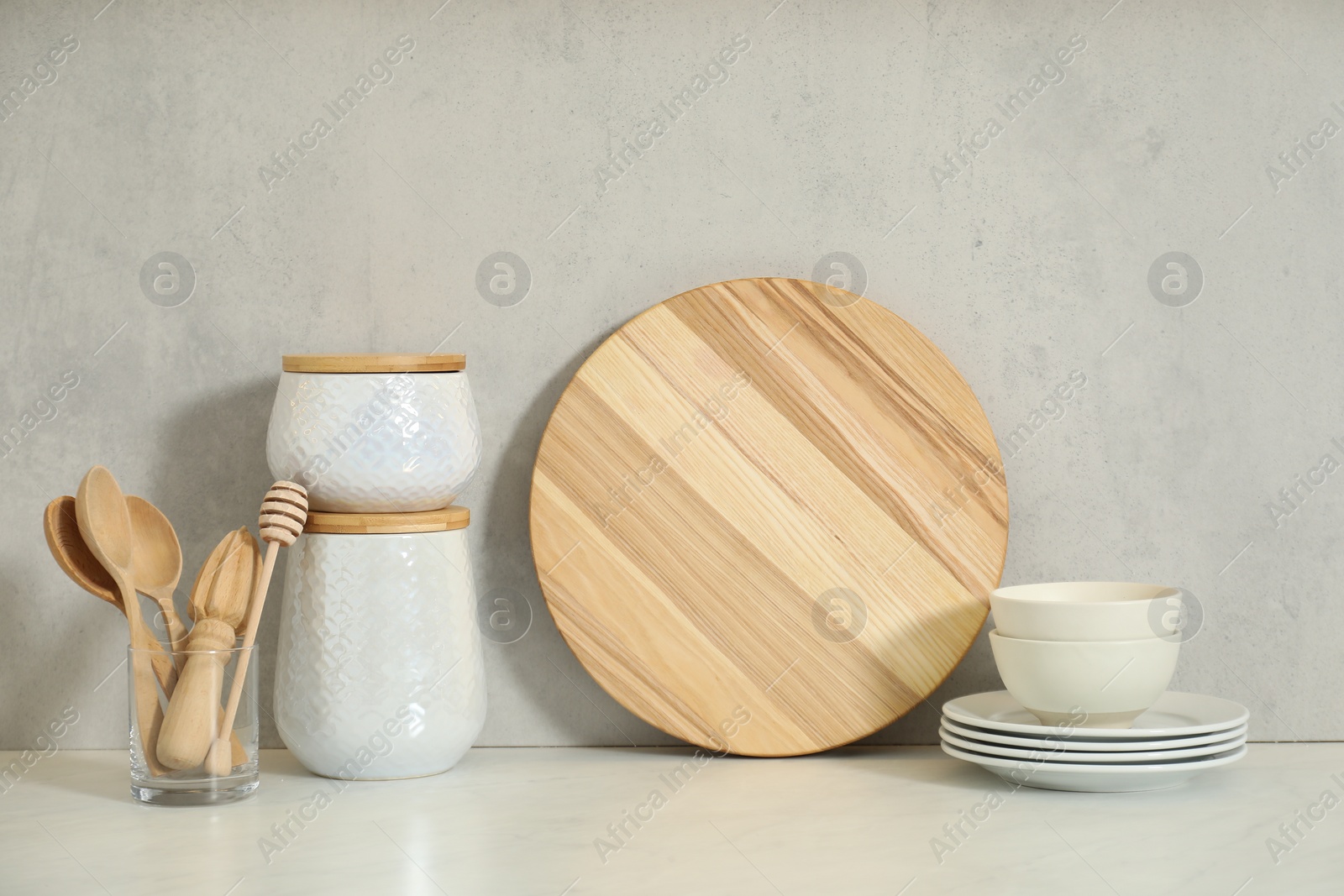 Photo of Wooden cutting board, other cooking utensils and dishware on white table near light wall