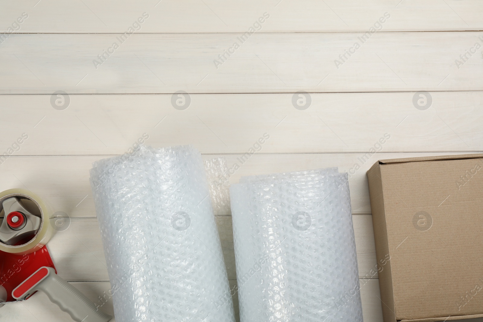 Photo of Bubble wrap rolls, cardboard box and tape dispenser on white wooden background, flat lay. Space for text