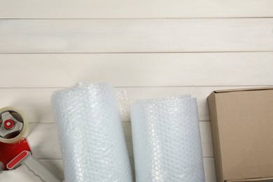 Photo of Bubble wrap rolls, cardboard box and tape dispenser on white wooden background, flat lay. Space for text