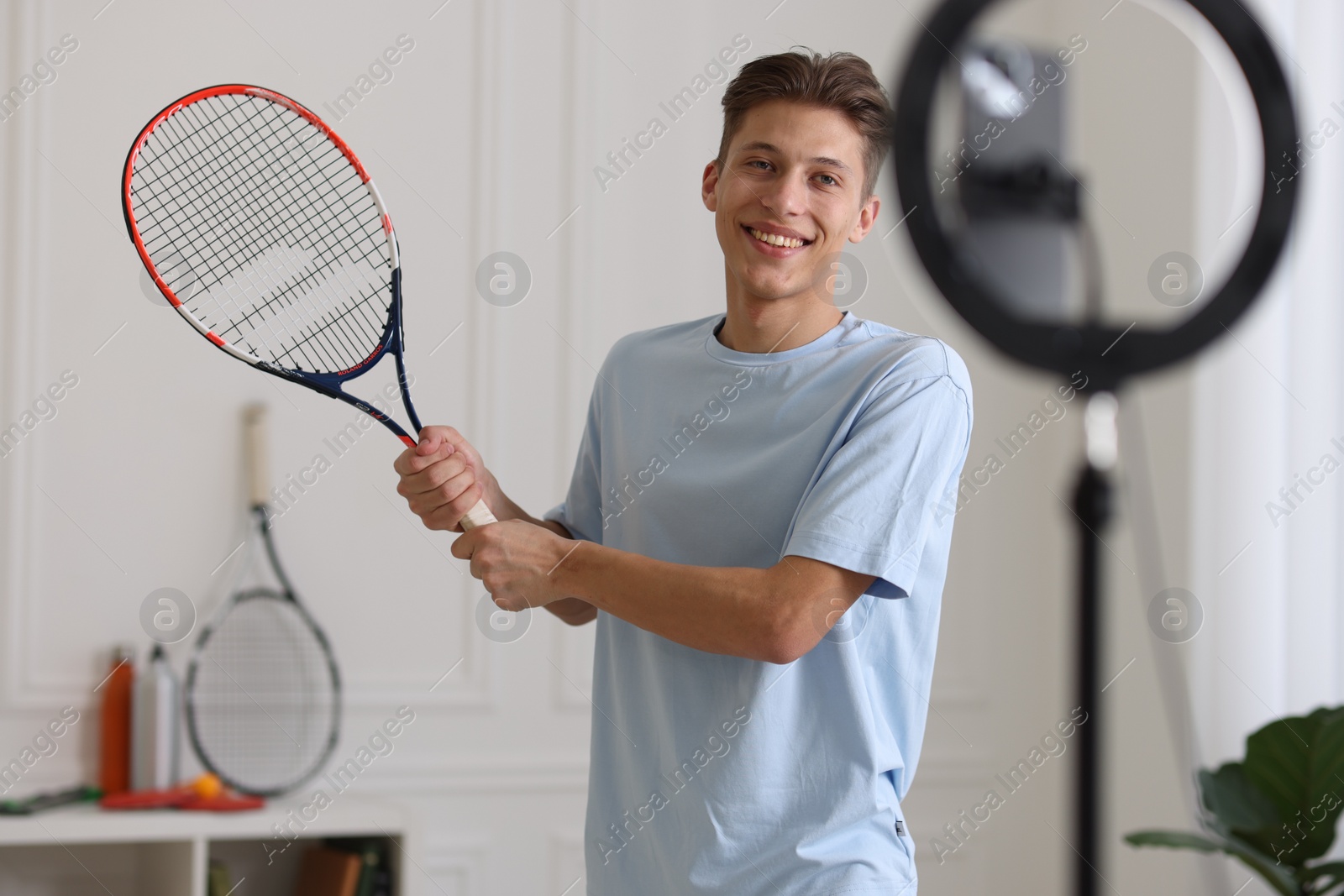 Photo of Smiling sports blogger holding tennis racket while streaming online fitness lesson at home