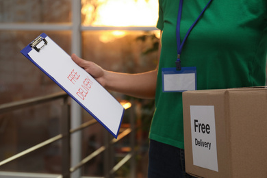 Photo of Courier holding parcel with sticker "Free Delivery" and clipboard indoors, closeup