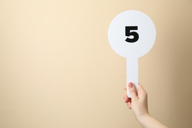 Woman holding auction paddle with number 5 on beige background, closeup. Space for text