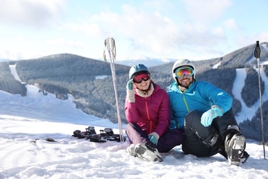 Happy couple with ski equipment sitting on snowy hill in mountains, space for text. Winter vacation