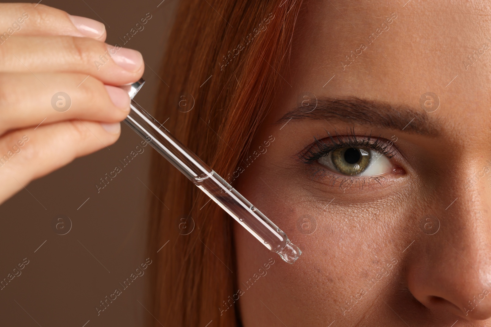 Photo of Beautiful young woman applying cosmetic serum onto her face on brown background, closeup