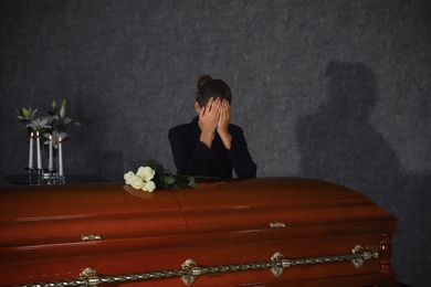 Photo of Sad young woman mourning near casket with white roses in funeral home