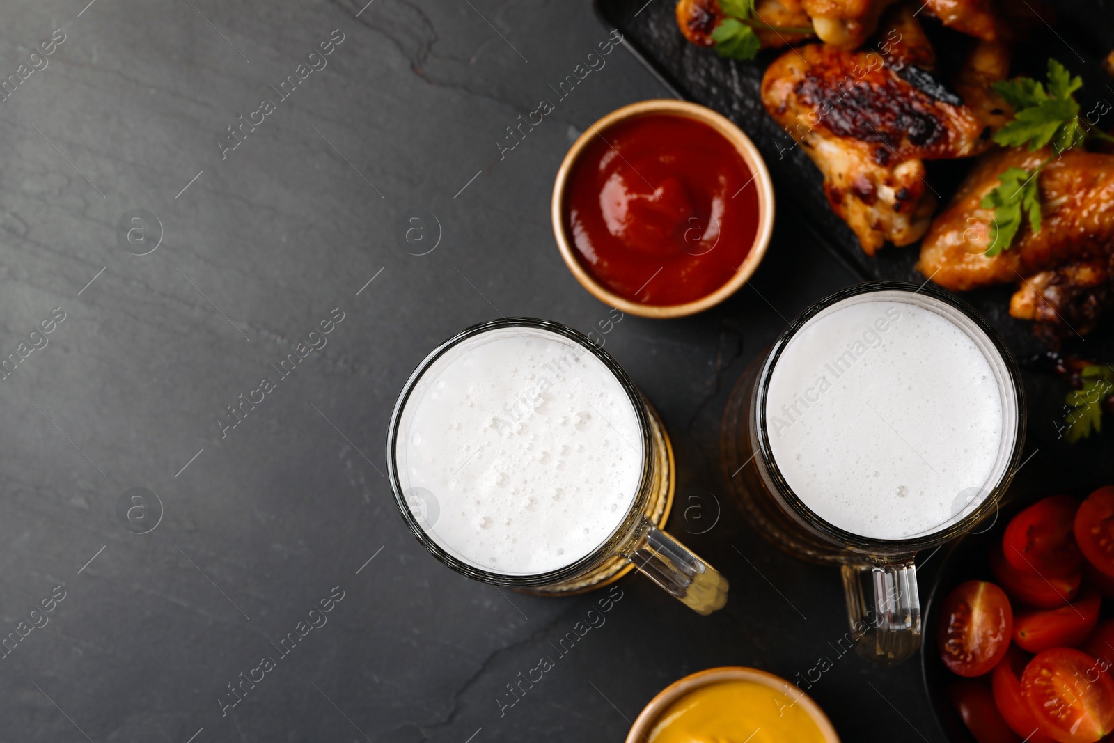 Photo of Mugs with beer, delicious baked chicken wings and sauces on dark grey table, flat lay. Space for text